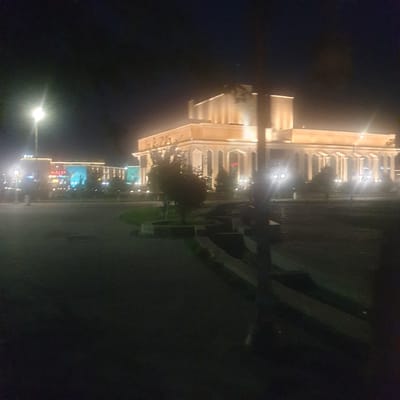 The image depicts a nighttime scene in Turkistan, Kazakhstan. A brightly lit building with an ornate facade stands prominently on the left. Moving further left along the lit street are several establishments with signs and displays, including one that says "HALS". The street stretches into the distance, illuminated by streetlights. A dark expanse of road occupies the right side of the image, with some blurred shapes suggesting benches or other roadside furniture. The sky above is dark. The overall impression is of a well-lit urban environment at night.