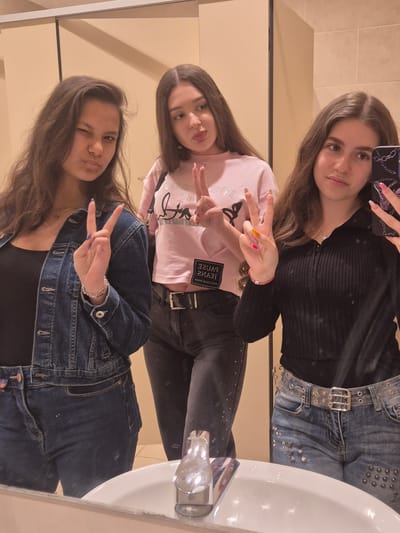 The original media file is a mirror selfie taken by three young women in what appears to be a bathroom setting, specifically in Zagore, Bulgaria. The image is rotated 90 degrees counter-clockwise.
When viewed upright, the scene features three young women posing for the camera.
The woman on the left has long, straight, dark brown hair and looks directly at the camera with a soft smile. She wears a black ribbed long-sleeved top and light blue jeans featuring silver studs and a distinctive clear belt with a silver buckle. She is holding a smartphone with a black case adorned with a silver chain pattern, which is being used to capture the image. Her nails are painted a light pink.
The middle woman has long, wavy, dark brown hair and is making a peace sign with her right hand, showcasing multi-colored nails (orange, pink, and brown/nude). She is wearing a light pink short-sleeved top on which mirrored text "PAUSE JEANS" is visible. She also wears dark-colored pants and a black belt. A thin red bracelet with a silver charm is on her wrist, and she looks towards the camera with a slight pout and gentle smile.
The woman on the right has long, dark brown hair and strikes a playful pose, winking and pouting while flashing a peace sign with her left hand. Her nails are painted a dark blue or purple. She is dressed in a black top underneath a blue denim jacket and dark blue jeans. A silver chain bracelet is visible on her left wrist.
The setting appears to be a bathroom, indicated by a white sink visible on the right side of the frame and light-colored tiled walls in the background. The overall mood is casual and friendly, with the subjects displaying expressions of enjoyment and playfulness.