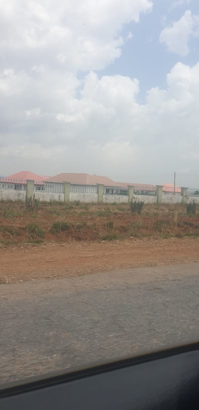 The image presents an outdoor scene in what appears to be Hoss, Nigeria, under a partly cloudy sky. The perspective is from ground level, likely from or near a road.
In the foreground, on the left side of the frame, there is a dark grey, paved surface, resembling an asphalt road. Adjacent to this road is a strip of reddish-brown, unpaved earth, sparsely covered with dry, light-colored grass and some small green shrubs.
Further back, a row of multiple buildings stretches across the mid-ground. These structures have uniform designs with distinctive reddish-brown, possibly corrugated metal, roofs. The walls appear to be made of light-colored concrete or block, featuring numerous window openings, some fitted with horizontal grates or frames. The buildings suggest a planned development or residential/commercial area.
The upper portion of the image is dominated by the sky, filled with scattered white and light grey cumulus clouds, with patches of lighter blue visible between them. The overall lighting indicates it is daytime, possibly an overcast or partly cloudy day.
No people or animals are visible, and there is no specific activity taking place. The scene is still and observational. The rightmost edge of the original image shows a dark, indistinct area, possibly a part of a car's interior or window frame from where the photo was taken. No visible text is present in the image.