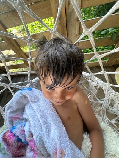 A young child, likely a boy, rests in a white macrame hammock suspended on a wooden structure. The child is partially covered by a white towel with colorful floral patterns. Sunlight casts shadows, suggesting it is daytime, and the lush green foliage visible through the wooden slats implies an outdoor setting. The child's dark, wet hair and relaxed posture suggest they may have recently been swimming or playing outdoors. They are looking up and to the side, with a neutral or slightly curious expression. The hammock is made of thick white rope, intricately knotted to form a net. The wooden structure appears rustic, possibly part of a treehouse or outdoor play area. No visible text is present in the image.
