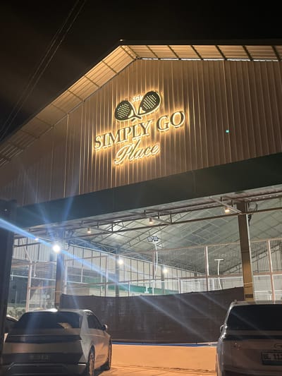 The image depicts the entrance to an indoor sports facility, likely a padel or tennis court, at night. A neon sign reads "SIMPLY GO Place" with two stylized rackets. The facility appears to have a corrugated metal roof and is illuminated by artificial lights, casting a warm glow on the signage. Outside the entrance, a white SUV is parked. The overall scene suggests a modern recreational space, potentially a popular spot for evening sports activities in Bayu, Indonesia. The lighting conditions and the presence of cars indicate it's nighttime. The location context of Bayu, Indonesia, helps place the scene.