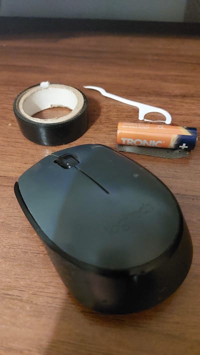 The image depicts a wooden table surface with several objects. In the foreground, a dark gray computer mouse is positioned. To the upper left of the mouse is a roll of black electrical tape. To the right of the tape, there is a white plastic dental floss pick. Adjacent to the floss pick is an orange and blue battery labeled "TRONIC" and a piece of dark gray material beneath it. The background is dark and out of focus. There is no visible indication of the city of Dubrovytsia, Ukraine, or any urban environment. The scene is static and appears to be a still life arrangement of everyday objects on a table.