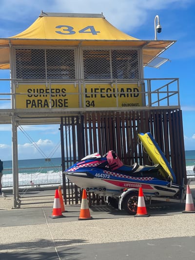 A bright yellow lifeguard tower numbered "34" stands against a blue sky with scattered clouds. The tower has "SURFERS PARADISE" and "CITY OF GOLDCOAST" signage. In front of the tower, a blue and red jet ski is parked on a trailer. The jet ski has "LIFEGUARD" and a number "464372" printed on its side. Orange and white traffic cones are placed on the ground around the jet ski. The background shows a sandy beach and the ocean with waves.