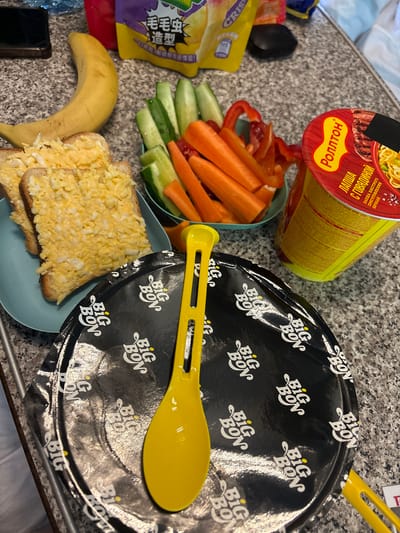 The image depicts a tabletop scene with various food items and a plastic spoon. In the foreground, a black paper plate with a repeating "Big Bon" logo is visible, along with a bright yellow plastic spoon resting on it. To the left, there are two slices of bread topped with a mixture that appears to be egg salad. Behind the bread, a banana is positioned. Further back, a bowl contains cut vegetables, including cucumber sticks and carrot sticks, along with red bell pepper slices. A red cup of instant noodles, identified by the "Rollton" brand, is also present. In the background, a yellow snack bag with Chinese characters and the word "CRISP" is visible, along with a dark object that could be a phone. The scene appears to be set on a speckled countertop. The location is identified as Nizhnyaya Kosa, Russia.