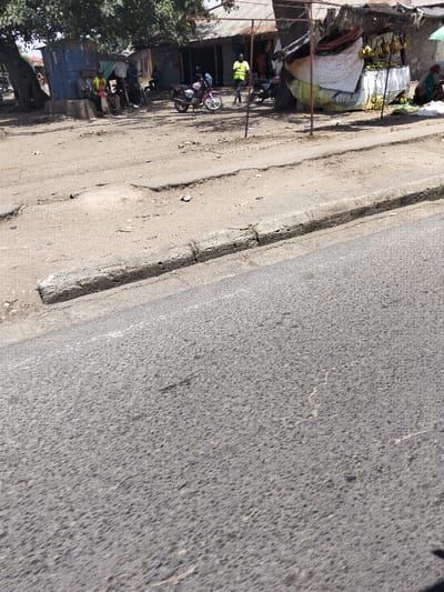 The image captures a roadside scene in Mkomani ward, Kenya, under daylight conditions. The foreground consists of an asphalt road exhibiting minor wear, bordered by a weathered concrete curb. Beyond the curb, an unpaved, dusty dirt and gravel shoulder contains small debris and uneven terrain.
In the midground, a cluster of informal structures and individuals is visible. On the far left, a large tree with green foliage shades a blue corrugated metal shack. Adjacent to this, at least three men are seated on a low structure or bench; one wears a yellow-green vest. Near the center, a red and silver motorcycle is parked on the dirt ground, facing left. Standing to its right is a man in a bright yellow-green shirt and dark trousers.
To the right of the center, a makeshift fruit stall is erected, featuring a framework of poles draped with white and yellow fabric or tarpaulin. Bunches of bananas are suspended for display, alongside what appears to be green produce. A person, possibly a woman, is seated on the ground next to bags at the base of this stall. Behind these elements, additional informal buildings with corrugated metal roofs are discernible. No active interactions are clearly observed among the individuals.