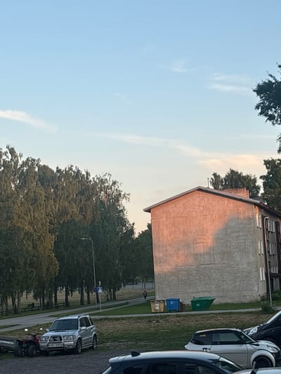 The image shows an outdoor scene in Tapa, Estonia. The sky is a pale blue with a few wispy clouds, suggesting it is daytime, likely late afternoon or early evening due to the warm light on the building.
The main subjects are a weathered, light-colored building with a sloping roof and a section of a paved area with parked vehicles. The building appears to be residential or perhaps a commercial property, with visible windows and a textured facade. A section of the building is illuminated by warm, golden light, casting a shadow across its surface. In front of the building, there are several parked cars. One car, a silver SUV, is prominently featured, its front grille and headlights visible. Behind it, other vehicles, including a dark-colored car and parts of what appear to be other vehicles, are visible.
To the left of the building, a dense line of green trees creates a natural backdrop. Another section of trees is visible further down the street. On the paved area near the building, there are three large trash bins: one green, one blue, and one yellow. A street lamp is visible in the distance, along with a pedestrian crossing sign. A small red and black all-terrain vehicle (ATV) is parked in front of the silver SUV.
The scene is calm and appears to be a typical residential or urban environment. There is no visible text in the image.