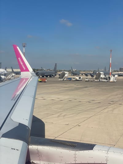 The image depicts an airport tarmac with several aircraft, including military-style planes and a commercial aircraft wing in the foreground. The wing in the foreground is white with a pink tip and the "wizzair.com" logo is visible. There are also passenger stairs and service vehicles on the tarmac. The background shows a clear blue sky with some clouds and distant buildings. This scene is located at Ben Gurion Airport, near the city of Yagel, Israel.