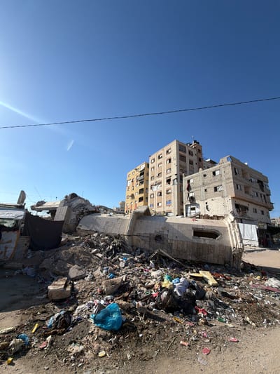 In the city of Jabalia, Palestinian Territories, a scene of destruction is depicted under a clear blue sky. In the foreground, a large pile of rubble and debris dominates the landscape. This rubble consists of shattered concrete, twisted metal, and various fragments of building materials, indicative of significant structural damage. Scattered amongst the debris are numerous items of refuse, including plastic bags, scattered garbage, and what appear to be discarded household items, creating a scene of disarray and neglect.
Behind the immediate foreground, several damaged multi-story buildings stand. The building on the far right exhibits numerous holes and impact marks on its facade, with some windows broken or missing. Other buildings in the background also show signs of damage, with some appearing partially intact but weathered. A banner with Arabic text is visible hanging on a structure to the left of the rubble. The overall environment suggests a community that has experienced conflict and subsequent damage to its infrastructure.
