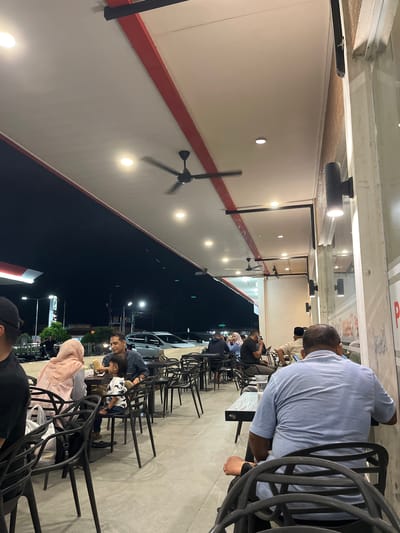 The image captures a nighttime scene within a semi-outdoor or covered dining area in Meunasahceubrek, Indonesia. Numerous individuals, primarily adults and some children, are seated at dark tables on black, open-backed chairs. The atmosphere appears casual and social, with people engaged in conversation, looking at their phones, or tending to children.

The dining area features a light-colored (possibly white or cream) ceiling with a prominent red horizontal stripe running along the upper wall. Recessed circular lights and track lighting fixtures illuminate the space, casting a bright glow. Several ceiling fans are visible, some in motion, indicating an effort to keep the area cool. Beyond the covered area, the sky is dark, characteristic of nighttime, with faint streetlights visible in the distance. Parked cars can be seen outside the perimeter of the dining space.

On the ground near one of the tables, a small white item, possibly a baby carrier or bag, is visible. The attire of some individuals, such as a woman wearing a hijab, provides cultural cues consistent with the Indonesian setting. People generally seem relaxed, enjoying their evening.

Visible text includes a red "P" on a white background, likely part of a sign, seen through a glass partition above one of the diners. Other indistinct white and red text is also visible on signs in the background.