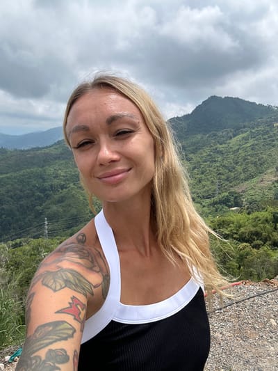 A woman with blonde hair and tattoos on her left arm takes a selfie outdoors. She is wearing a black tank top with a white trim. She is smiling at the camera. The background shows a lush green mountain range under a cloudy sky. The location is identified as Nam Hải, Vietnam.