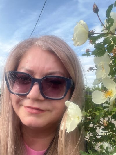 A selfie-style portrait features a blonde woman outdoors in Shud'ya, Russia, under a clear, bright blue sky with sparse white clouds. The image is rotated, showing the woman's head tilted to her right, with her long, light blonde hair flowing downwards and to her left. She wears dark-framed sunglasses that cover her eyes, reflecting a reddish tint. Her lips are slightly parted, revealing a subtle, relaxed expression, and she has light pink lipstick on. A pink garment is visible on her right shoulder and chest.
Overhead, from the viewer's perspective, are branches adorned with white or very pale yellow flowers, resembling wild roses, interspersed with green leaves. Some flowers are fully bloomed, showcasing yellow stamens, while others are still in bud form, and a few appear withered. These flowers create a natural, blooming canopy in the upper portion of the frame. In the top left, a dark, thin wire or power line stretches across the blue sky. The bright, natural lighting indicates it is daytime, likely morning or afternoon, with pleasant, sunny weather. There is no visible text in the image.