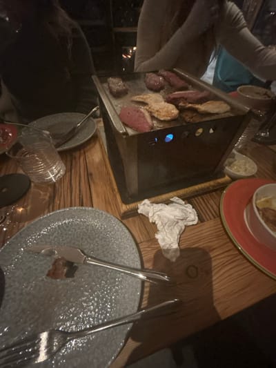 A wooden table displays various dining elements. A charcoal grill features partially cooked meat. A plate with a red rim holds an indistinct food item. A crumpled white napkin sits next to the grill. A textured gray plate hosts a knife and fork with a morsel of food. To the lower left, overturned drinkware adds to the scene.