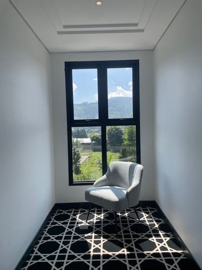 This image depicts an indoor scene within a building located in Kayuambon, Indonesia. The room features plain white walls and a white ceiling with a recessed rectangular architectural detail and a single, warm-toned circular recessed light fixture. A large, black-framed window occupies the central portion of the back wall, divided into multiple glass panes. A light grey upholstered armchair with slender black metal legs is positioned on a black carpet that covers the floor. The carpet exhibits a repeating geometric pattern of white intersecting lines, creating circular and star-like motifs. Sunlight streams through the window, casting pronounced shadows of the window frame and the armchair onto the patterned carpet. Through the window, a clear blue sky with scattered white clouds is visible above a lush, green, hilly landscape. The outdoor view includes numerous trees, some visible structures, and what appears to be terraced terrain in the background. No individuals are present in the scene.