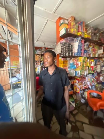 The image captures a scene inside a small, brightly lit shop, likely a convenience store or a local market stall, located in Akowonjo, Nigeria. The shop is densely stocked with various products, primarily packaged goods like snacks, beverages, and household items, arranged on shelves that line the walls. An orange plastic chair is visible in the foreground, suggesting a waiting area or customer seating. Two individuals are present. One, a young man wearing a black short-sleeved shirt and dark trousers, is standing in the center of the shop, facing left and appearing to be in conversation. Another person, partially visible on the left side of the frame, is wearing a blue shirt and is positioned near a barred entrance, also facing towards the shop interior. The overall impression is that of a busy retail environment, possibly during a transaction or interaction between the two individuals. The lighting is somewhat harsh, creating strong shadows on the floor, which is tiled with a patterned design. The back wall of the shop is painted a vibrant orange, contrasting with the white ceiling.