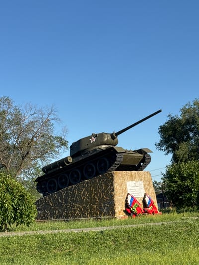 The image shows a T-34 Soviet tank monument in the city of Zimovniki, Russia, under a clear blue sky. The tank is positioned on a stone pedestal adorned with two wreaths, one on each side, featuring red, white, and blue ribbons and red flowers. A plaque is affixed to the pedestal above the wreaths. The monument is surrounded by green grass and trees, with a light-colored pathway visible in the foreground. The monument is a likely tribute to military history, given the presence of the tank and the wreaths.