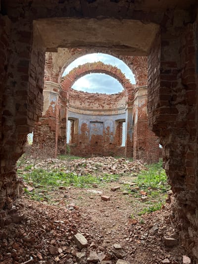 The image presents the interior of a severely ruined brick building, likely an abandoned church or a large historical structure in Staroe Rakomo, Russia. The photograph is taken from within a dark, narrow archway, looking into a larger, dilapidated space.
The most striking feature is a massive, partially collapsed circular opening in what appears to be the former dome or ceiling, through which a bright, overcast sky is visible. Below this, the curved interior walls are constructed from reddish-brown brick, with much of the original plaster and decorative finishes having eroded or fallen away, revealing the rough brickwork beneath. Traces of architectural elements, such as cornices, moldings, and pilasters, are still discernible on the crumbling surfaces. Several rectangular window openings are set into these walls; one retains what looks like a metal grate, while others are open to the outside.
The ground within the ruin is uneven and covered with a thick layer of debris, including broken bricks, stones, and earth, suggesting a long period of decay. Patches of vibrant green weeds and other vegetation grow vigorously amidst the rubble, particularly on the right side of the frame, indicating nature's reclamation of the abandoned space. The lighting is natural, with diffused light from the overcast sky illuminating the interior, while the foreground and the entry archway are cast in deeper shadow. There are no people visible in the scene, and no discernible text. The image evokes a sense of decay, history, and the relentless passage of time.