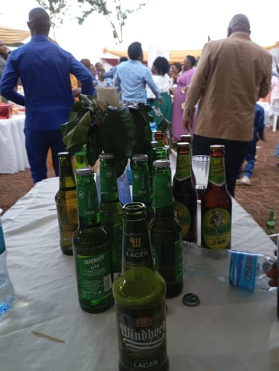 This media file captures an outdoor social gathering or event in what appears to be Shimbi Mashariki, Tanzania, indicated by the local beer brands. The foreground is dominated by a white tablecloth covering a table laden with numerous beverage bottles.
**Subjects:** Several individuals are visible in the background, mostly out of focus. A man in a blue shirt stands with his back to the camera on the left, while another man in a light brown jacket is seen from behind on the right. Other guests, including women in dresses, are scattered in the mid-background, mingling. A floral arrangement featuring a prominent white rose is placed centrally on the table.
**Objects:** The table holds a variety of empty and possibly some full beer bottles. Brands clearly visible include "Serengeti Lite", "Serengeti Lemon", and "Windhoek Premium Lager". Some bottle caps bear the "18+" age restriction symbol. A clear plastic water bottle with a blue cap is on the left, and a crushed plastic bottle with "MAJI ORO" written on its blue label is on the right. A single toothpick lies on the tablecloth. In the background, white chairs and tables with white coverings are set up, and a yellow or gold tent structure is partially visible. Red gift boxes are seen on a table to the left.
**Scene/Setting:** The event takes place outdoors on what appears to be a dirt or sandy ground. The sky suggests daytime, possibly overcast, providing soft, diffused lighting. The setup with multiple tables and chairs indicates a prepared event space.
**Activity or event taking place:** The scene depicts a social gathering, possibly a celebration, where people are mingling and enjoying beverages. The presence of many consumed bottles suggests an ongoing or recently concluded celebratory meal or party.
**Notable details:** The soft natural light suggests it's daytime. The specific beer brands, Serengeti and Windhoek, are common in East and Southern Africa, aligning with the Tanzanian location context. People in the background appear relaxed, indicating a pleasant atmosphere.
**Visible text:** Text visible on bottles includes "SERENGETI LITE", "4.0% 330ml", "SERENGETI LEMON", "Freedon in the flavor" (partially), "WINDHOEK PREMIUM LAGER", and "PREMIUM LAGER". Bottle caps show "18+". A crushed water bottle displays "MAJI ORO".