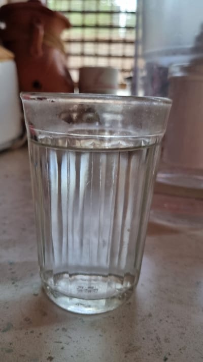 A close-up shot shows a clear glass with vertical fluting, filled with a clear liquid, sitting on a speckled surface. The glass is positioned in the foreground, with its rim slightly fogged. In the background, out of focus, a brown, amorphous object rests to the left, and a cylindrical, translucent container is visible to the right. Behind these, a window with horizontal blinds is apparent, allowing diffused light to enter. The overall scene appears to be indoors, potentially in a kitchen or dining area. The location is identified as Солнечная Поляна-3 Снт, Russia.