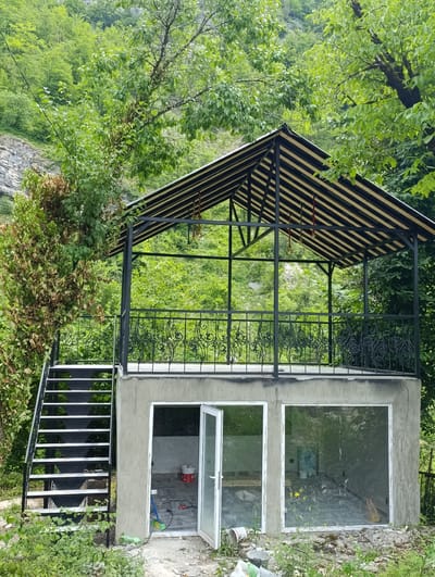 The image shows a two-level structure with an outdoor gazebo on the upper level and a room with windows and a door on the lower level. The gazebo has a black and yellow striped roof and a black metal railing. An external metal staircase provides access to the upper level. The lower level room has large windows and a door that is ajar, revealing construction materials inside. The structure is situated in a lush, green environment with trees and foliage surrounding it. The background consists of a forested hillside and rocky terrain, consistent with the mountainous landscape of Adjara, Georgia. There are no people visible in the image.