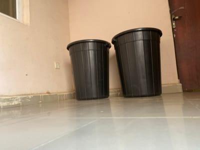 Two black plastic trash cans are positioned on a light-colored tiled floor in front of a cream-colored wall. The trash can on the left is slightly closer to the viewer, while the one on the right is further back and to the right. Both trash cans have a ribbed exterior and a wide rim. A dark brown door with a silver handle is visible to the far right of the frame, partially obscured by the trash can on the right. A window frame with a light-colored edge is visible in the upper left corner of the image. The base of the wall shows signs of wear, with a lighter colored border and some streaks or drips. The scene appears to be indoors, possibly in a hallway or room with a door and a window. The overall lighting is neutral.