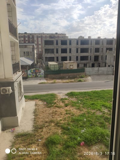 An eye-level, medium shot captures an unfinished concrete building in the city of Charjuy, Uzbekistan, against a cloudy sky. The building, constructed with grey concrete blocks, features numerous dark, empty window frames. A temporary green metal fence encloses part of the construction site, with a small, light-colored utility box situated near the fence. To the left of the construction site, a smaller, older building with a grey roof is visible, adorned with a colorful mural depicting cartoon characters and a tree, with the word "KAMALA" prominently displayed. In the foreground, a paved road runs horizontally across the frame, bordered by a strip of dry, patchy grass with scattered litter. A modern apartment building with a black metal balcony is partially visible on the far left. An outdoor air conditioning unit is mounted on the grey exterior wall of this building. The overall scene suggests ongoing construction in an urban environment, with signs of wear and tear in the surrounding public spaces.