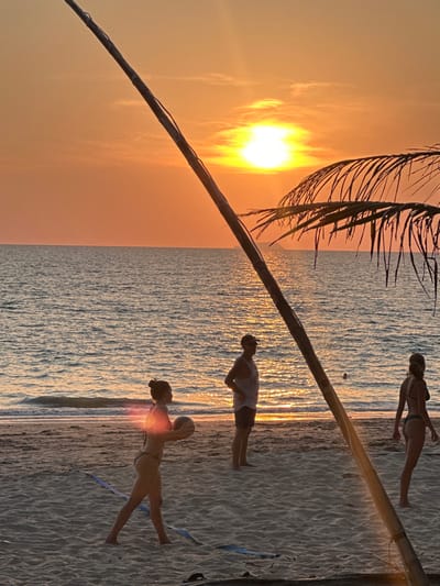 An outdoor scene captures a sunset over a sandy beach in Phra Ae, Thailand. The sun, a bright yellow-orange sphere, is low on the horizon, dominating the sky and reflecting intensely on the ocean's surface, where small waves are visible. A prominent wooden pole, likely part of a beach volleyball net, extends diagonally across the frame from the lower left to the upper right. A palm frond is visible in the upper right quadrant.
Three individuals are present on the beach. In the foreground, a female, silhouetted by the backlight and partially obscured by a lens flare, wears a dark two-piece swimsuit and holds a light-colored volleyball with both hands, positioned in front of her. She stands in a ready stance, facing towards the right side of the frame. To her right and slightly behind, a male individual, also partially silhouetted, wears a light tank top, dark shorts, and a cap, standing with his hands possibly at his sides, facing left. Further to the right, another female individual in a dark two-piece swimsuit stands facing left, with her back mostly towards the viewer. Blue court lines are visible on the sand in the foreground. The scene depicts recreational activity on the beach during sunset.