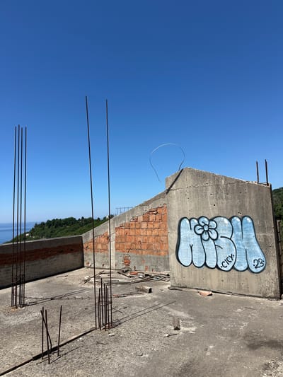 The image shows a building under construction in Gornji Katun, Montenegro. The walls are a mix of concrete and brick. Graffiti art is visible on the concrete wall. Rebar extends upward from the concrete platform, suggesting the structure is incomplete. The sky is a clear blue. In the background, there are trees and a glimpse of the sea.