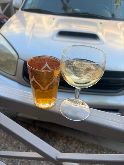 An outdoor scene in Cagnes-sur-Mer, France, depicts two glasses resting on a gray metal railing. To the left, an amber-tinted glass tumbler, adorned with intricate gold and pink-toned patterns, contains an amber liquid. To the right, a clear stemmed wine goblet, tilted slightly, holds a pale yellow, slightly effervescent liquid. Both glasses are positioned on a horizontal bar of the railing, with the ground surface below them indistinct. In the background, a silver-colored Toyota RAV4 SUV is parked, with its hood and upper front grille visible, rendered out of focus. Sunlight reflects off the vehicle's metallic surface, indicating daylight conditions. No individuals are present in the image.