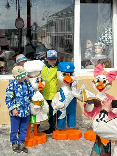 The scene takes place in front of a store window in what appears to be the city of Arzamas, Russia. Two young boys are interacting with large, colorful, folk art-style figures of cartoonish ducks, which are displayed on the sidewalk.

The boy wearing a blue and white patterned jacket and blue pants is lying down and appears to be hugging the duck figure dressed as a chef, complete with a white hat. The other boy, wearing a light green jacket and a blue baseball cap, is lying down and appears to be hugging the duck figure dressed as a mechanic, wearing a blue cap with the Volkswagen logo and holding a large wrench.

There is a third duck figure, dressed as a lady with a pink bow and eyeglasses and holding a book, being photographed by an adult partially visible on the right. These are lined up in front of an exterior wall, each duck figure is attached to a set of orange "legs" or supports.

The store window behind the ducks reflects the street scene and displays mannequins wearing hats and headscarves. The background shows buildings and street furniture, characteristic of an urban setting. The light suggests it is daytime.