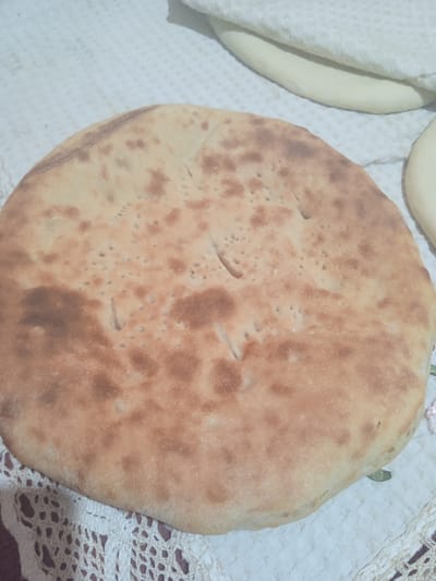 The image displays a close-up of a round, flatbread on a white, patterned tablecloth. The flatbread is golden brown with darker browned spots and numerous small indentations across its surface. The bread appears to be freshly baked. In the background, slightly out of focus, are additional layers of what appear to be dough or more flatbread, covered by a white textured fabric. A sliver of another flatbread is visible on the right edge of the frame. The scene is devoid of any human presence or explicit geographical markers that would pinpoint a location in the city of Теңги, Kyrgyzstan.
