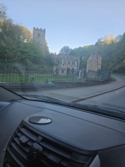 The image presents a picturesque view of a historic landscape, captured from the interior of a vehicle. The lower part of the frame reveals the dark grey dashboard and a portion of the windshield, including an air vent and a circular control, indicating the perspective of a passenger or driver.
In the mid-ground, a dark tarmac road, appearing damp or wet, curves gently from the left towards the right. Alongside the road, a neatly mown green lawn with distinct stripes extends towards a series of ancient stone buildings.
Two prominent historical structures dominate the scene. On the right, a large, multi-story stone building features numerous arched windows and intricate stonework, resembling an old church, abbey, or grand estate. To its left and slightly further back, a sturdy, square stone tower with battlements and a single visible window stands, partially framed by trees. The architectural style suggests a significant historical presence, characteristic of the United Kingdom.
Beyond the buildings and rising up a gentle incline, a dense canopy of green trees covers the hillside, with some deciduous trees appearing to have lighter or sparser foliage, possibly indicating early spring or late autumn. Above the trees and buildings, a clear, bright blue sky completes the tranquil setting.
The image captures a static moment during daylight hours, likely morning or late afternoon, given the clear sky and even lighting. The wet road, juxtaposed with the clear sky, suggests recent rain has passed, leaving the landscape refreshed. No specific activity is taking place other than the observation of the scenic view from the vehicle. There is no visible text within the image.