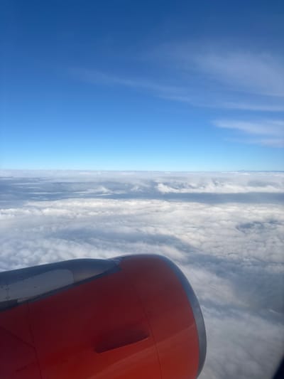The image shows an airplane view. A portion of the orange-colored engine is visible on the right side. The left side displays a clear blue sky, while the center shows layers of clouds. The location is not discernible from the image and thus, a relation to the city of Horsham, United Kingdom, cannot be established.
