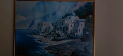 This is an oil painting depicting a coastal town with white buildings and a blue-domed church, nestled against a backdrop of steep, rocky hills or mountains. The scene is characterized by a strong use of blue and white, suggesting a bright, possibly Mediterranean, day. In the foreground, several boats are pulled up onto a pebbled beach, with gentle waves lapping at the shore. The architecture of the buildings is indicative of a Greek island, with the white walls and blue domes being a common feature. Given the architecture, the scene is likely located in a Greek coastal town. While the specific location within the Municipal Unit of Aegio is not definitively identifiable from the image alone, the style of architecture strongly suggests a Cycladic island aesthetic, which is not typical of Aegio itself, but common in many Greek islands. The painting is framed in a simple gold frame. There are no people clearly visible in the painting. The painting has a slightly blurry or soft focus quality, which is characteristic of the artistic style rather than an issue with the photograph.