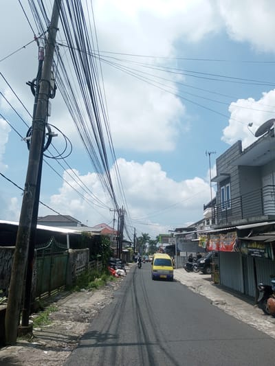 The image shows a street scene in Xayabury District, Laos. A yellow and blue van is traveling down a narrow, dark asphalt road. The left side of the road has a worn concrete curb, a dilapidated fence, and a utility pole heavily laden with bundled wires. The right side of the road has buildings, some with signage, and parked motorbikes. The sky is blue with scattered white clouds. The overall scene depicts a common street view in a developing urban area.