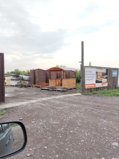 A full shot captures an outdoor storage yard on a cloudy day. In the foreground, the gravel-covered ground transitions to a concrete curb and then to a dirt path. To the left, a portion of a vehicle's side mirror and door is visible, reflecting the yard's contents. The midground is dominated by stacks of lumber, wooden structures resembling sheds, and covered materials on pallets. A large wooden billboard stands on the right side, displaying advertisements for construction materials, including "PENOPLEX." In the background, more wooden structures and trees are visible under an overcast sky.