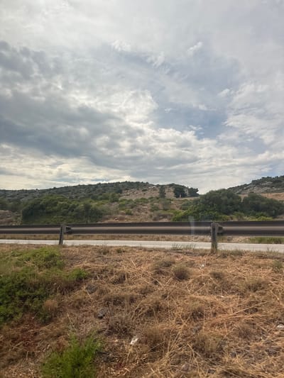 The image depicts a roadside view with a steep, vegetated hill on the left and a dry, grassy embankment on the right. A metal guardrail runs horizontally across the middle of the frame, separating the road from the embankment. The hill is covered with a mix of green trees and scrubby bushes, with rocky outcrops visible in places. The sky is filled with dramatic, grey and white clouds, suggesting overcast or stormy weather. The dry grass on the embankment is a light brown and yellow, indicating a dry season or arid environment. The scene appears to be a natural landscape, possibly a rural or mountainous area. There are no people or animals visible in the image. The time of day is difficult to determine precisely due to the cloud cover, but the lighting suggests it is daytime. The location context suggests this scene is near Salses-le-Château, France.
