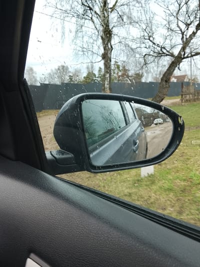 The image shows the view from inside a car, focusing on the driver-side rearview mirror. The mirror reflects a gray or silver car traveling on a dirt road. In the background of the mirror's reflection, a white car is visible in the distance. Outside the car window and in the mirror, the environment consists of bare trees, a gray fence, green grass, and some houses. The picture was taken in rainy weather, as evidenced by the raindrops on the window and mirror. Judging from the architectural style of the houses, and the bare trees and unpaved road conditions in the mirror's reflection, this scene is likely located in a rural or suburban area of Высокиничи, Russia.
