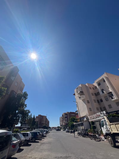 The image shows an urban street scene in Saâda, Morocco, under a clear, bright blue sky with a prominent sun flare. The street is lined with buildings of varying heights and architectural styles, ranging from modern apartment blocks to older residential structures. Parked cars are visible along the street. Vegetation, including trees, can be seen lining the street.