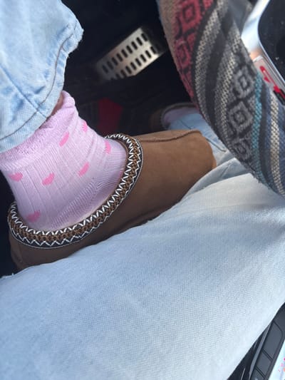Inside a vehicle, a person's lap is visible. The individual wears light-wash jeans. On one foot, a pink sock with small circles is paired with a brown slipper featuring a decorative trim. The driver's side is noticeable with a patterned steering wheel cover and a metallic pedal. The car's dashboard and controls are partially visible.