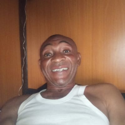 The image displays a close-up of an adult male with dark skin and a bald head. He wears a plain white tank top and exhibits a wide, open-mouthed smile, revealing his teeth, with his eyes directed forward. The background consists of light brown wooden paneling. A thin, dark vertical strip is visible along the far left side, with a wavy white cord running vertically alongside it. This scene is located in Koko, Nigeria.