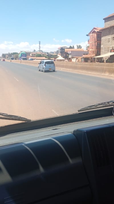 From inside a vehicle, a highway stretches into the distance under a clear, bright sky. A light blue compact car travels ahead in the same direction. Buildings and structures line the right side of the road, including a red-brick building. An antenna or cell tower is visible further in the distance. The vehicle's dashboard and a portion of the windshield are visible in the foreground.