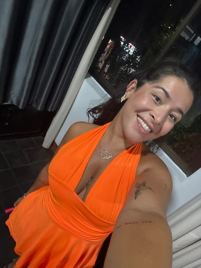 A woman with dark hair and brown eyes is taking a selfie. She is wearing an orange halter dress and gold jewelry. She has tattoos on her arm and chest. She is smiling at the camera. The background is dark, with some lights visible through a window. The scene appears to be indoors, possibly a hotel room or a restaurant, with a window overlooking a city at night. The location is not specified as Khan Daun Penh, Cambodia.