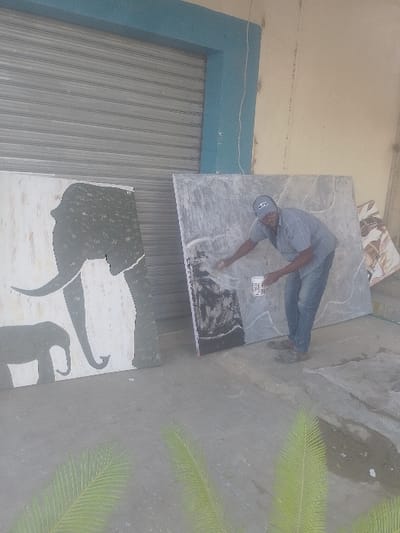 This image depicts an outdoor scene in Bamburi ward, Kenya. A male individual is shown actively working on a large rectangular panel. He is wearing a light blue short-sleeved collared shirt, blue denim jeans, dark closed-toe footwear, and a light-colored baseball cap with a dark logo. His right hand is applying a substance, possibly paint or sealant, to the lower-left portion of the panel, while his left hand holds a cylindrical white container. The panel he is working on is light grey with irregular dark grey and white vein-like patterns. To his immediate left, another large rectangular panel stands, featuring a prominent grey silhouette of an adult elephant and a smaller grey silhouette of a baby elephant against a white background. The background consists of a building with a closed grey corrugated metal roll-down security shutter, framed by a light blue header and vertical trim on the left. A plain, light cream-colored wall is visible to the right of the shutter. The ground is a flat, grey concrete surface exhibiting some darker, irregular patches. Two bright green palm fronds are partially visible in the lower foreground.