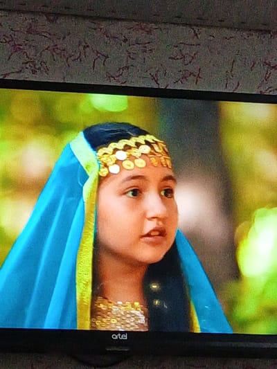 This image is a photograph taken in Suti, Uzbekistan, depicting a television screen displaying a close-up of a young girl. The girl, with dark hair, light complexion, and brown eyes, is positioned slightly off-center, looking towards the right of the frame with her mouth slightly open. She wears a vibrant teal-blue head covering edged with a lime-green trim, securing her dark hair. A gold-colored ornamental headpiece featuring multiple coin-like discs rests on her forehead. The top of a gold-sequined or elaborately embroidered garment is visible below her neck. The background on the screen is a blurred expanse of green foliage, suggesting an outdoor or natural setting. Below the depicted scene, the word "artel" is visible in white letters against the black lower bezel of the television screen. Above the screen, a portion of a patterned, possibly floral, wallpaper is visible.