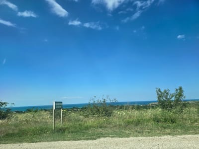 The image shows a wide, sunlit view of a grassy hillside sloping down towards a body of water, likely the sea, under a clear blue sky with scattered white clouds. In the foreground, a dirt road or path is visible. A green sign with white lettering stands on a wooden post in the middle ground. The sign is in Russian and appears to be a directional or informational marker. The hillside is covered in tall, green grass and wildflowers, with some scattered bushes and a small tree on the right side of the frame. In the distance, there are more trees and some buildings, suggesting a settlement. The overall impression is one of a natural, rural landscape on a bright, clear day. The provided crops confirm the presence of the sky, the sign, and the natural environment. The location is identified as Ullubiiaul, Russia.