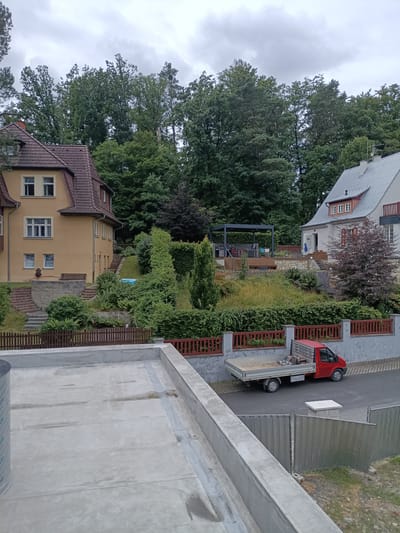 This media file depicts a residential street scene in Doksy, Czechia. The primary subjects are two houses, one painted yellow and the other white with a gray roof, nestled amongst lush green trees. In the foreground, a rooftop construction is visible, with concrete surfaces and some construction materials.
A red flatbed truck is parked on the asphalt road in front of a red wooden fence and a gray wall. The truck appears to be carrying some equipment. A small, blue swimming pool is visible in the garden area between the two houses. The scene suggests a daytime setting with overcast weather, as indicated by the gray sky. The overall mood is tranquil and everyday, with no discernible human activity beyond what can be inferred from the parked vehicle and residential setting. The location cues, such as the architecture of the houses and the surrounding vegetation, are consistent with a European setting. No visible text is present in the image.