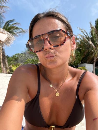 A woman is captured in a close-up selfie outdoors in a tropical setting, likely Thoddoo, Maldives. She is looking slightly upwards with a pouting expression, wearing large, tortoise-shell patterned sunglasses and a dark brown bikini top. Her skin is tanned and glistens with water droplets, suggesting she has recently been swimming or in contact with water. She is adorned with a gold chain necklace featuring a circular, coin-like pendant and a smaller heart charm, along with gold earrings. The background reveals a clear blue sky and lush green palm trees, indicating a bright, sunny day. A small, reddish tattoo is visible on her upper right arm. The overall scene conveys a relaxed, vacation atmosphere.