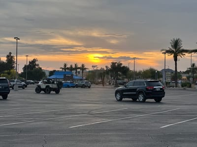 The image shows a parking lot in Phoenix, Arizona at sunset. In the background, signs for businesses, including TJ Maxx, Aldi, and Party City, are visible. An IHOP restaurant is also visible. Palm trees dot the landscape. There are several cars in the parking lot, including a black Jeep Grand Cherokee, and a white open-top jeep. The sky is filled with clouds illuminated by the setting sun.
