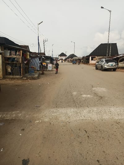 The image shows a street scene in Obromudu, Nigeria. The street is paved with asphalt, but it is cracked and dusty. There is one person walking down the street, and there are several buildings on either side of the road. There are two cars parked on the side of the street. There are a few streetlights present. The street is bordered by buildings and vegetation. The weather is cloudy. The overall scene is a typical street scene in a small town in Nigeria. The coordinates provided place the image in Obromudu, Nigeria.
