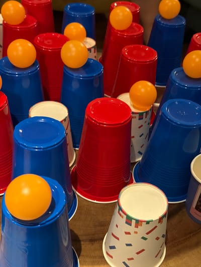 The image depicts a close-up view of numerous red and blue plastic cups, some of which have orange ping pong balls balanced on top. The cups are arranged in a somewhat chaotic but visually organized manner, with some stacked and others standing individually. One cup in the foreground features a colorful confetti-like pattern. The surface beneath the cups appears to be a brown, textured material, possibly wood or fabric. The lighting is moderate, casting subtle shadows. There are no people visible in the image. The scene is devoid of any specific landmarks or clues that would allow for identification of a location in the city of Heijen, Netherlands.